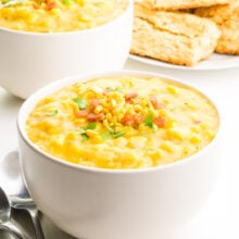A bowl of corn chowder has spoons next to it. There's another bowl of chowder and biscuits in the background.