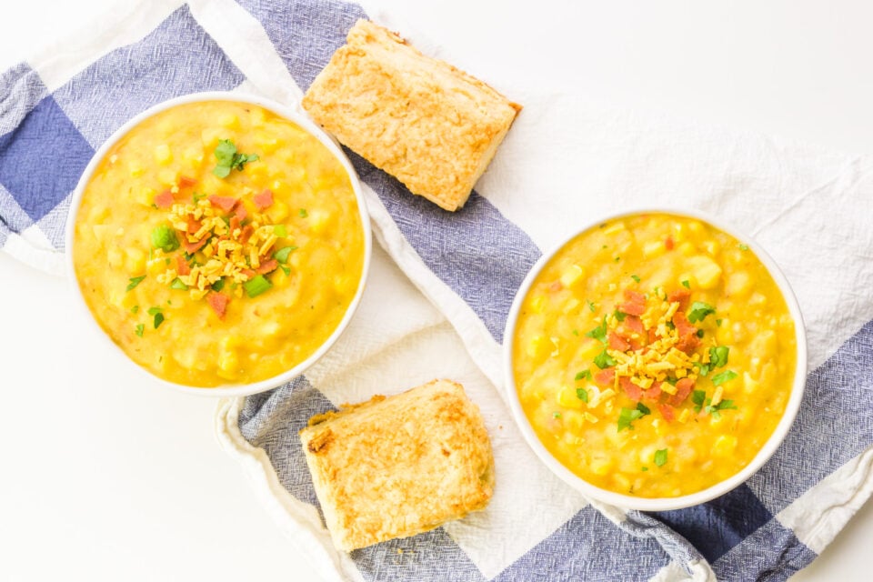 Looking down on two bowls of corn chowder sitting on a blue and white Kitchen towel. There are biscuits between them.