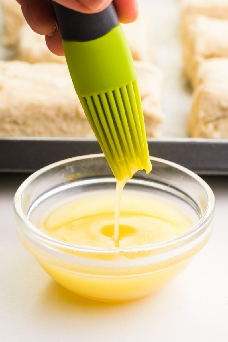 A silicone pastry brush is dipped in a bowl with vegan egg wash. A pan of pastries sits behind it.