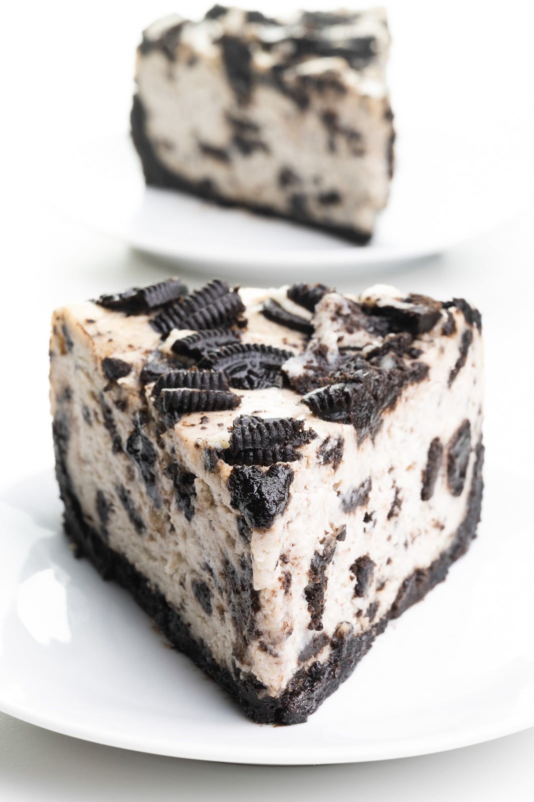A slice of vegan Oreo cheesecake sits on a plate. Another slice is behind it.