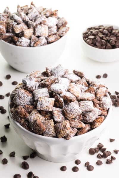 Two bowls are full of vegan puppy chow cereal treats. There are chocolate chips on the table and a bowl of chocolate chips in the background.