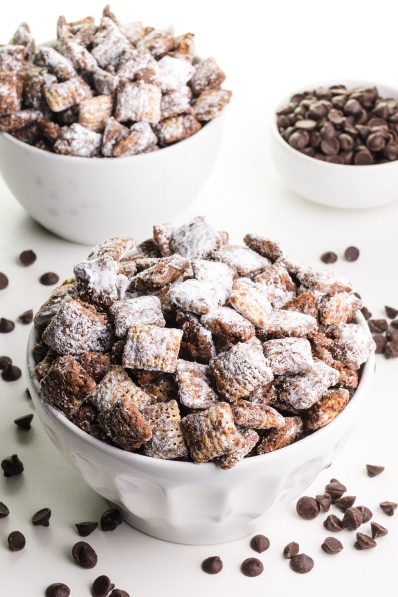 Two bowls are full of vegan puppy chow cereal treats. There are chocolate chips on the table and a bowl of chocolate chips in the background.