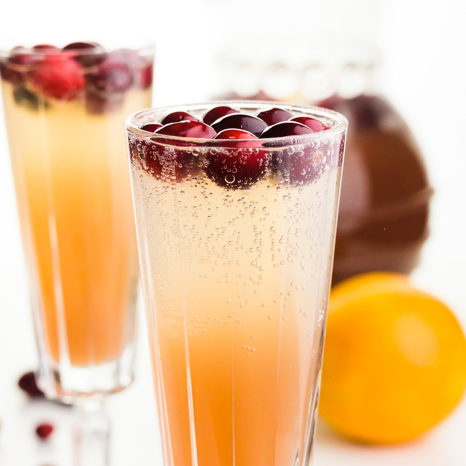 Two glasses of Christmas punch have fresh cranberries on top. There's a pitcher and an orange behind them.