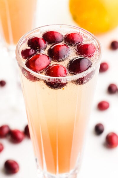 A close-up image of a glass of holiday punch shows fresh cranberries and sparkling water fizzes at the tope. There are fresh cranberries, an orange, and another glass in the background.