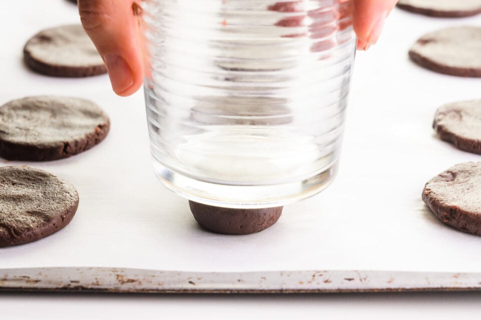 A hand holds a glass and is pressing cookie dough balls into round discs on a baking sheet lined with parchment paper.
