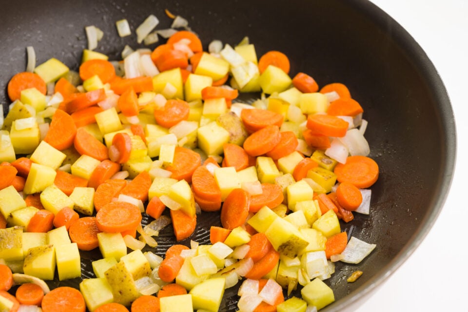 Chopped onions, carrots, and potatoes are cooking in a pan.