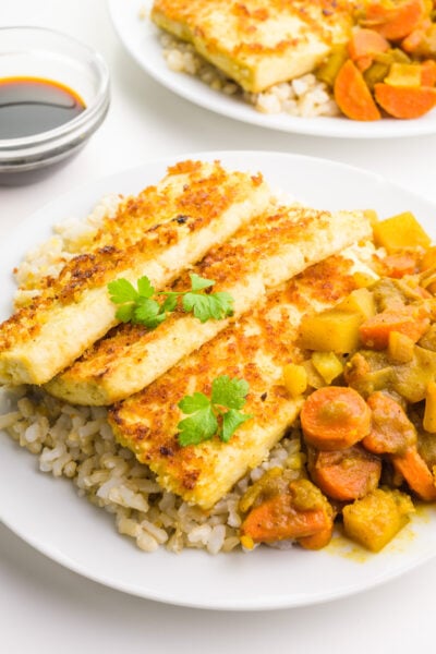 A plate has rice, fried tofu, and vegetarian katsu curry. There's a small bowl of soy sauce and another plate behind it.