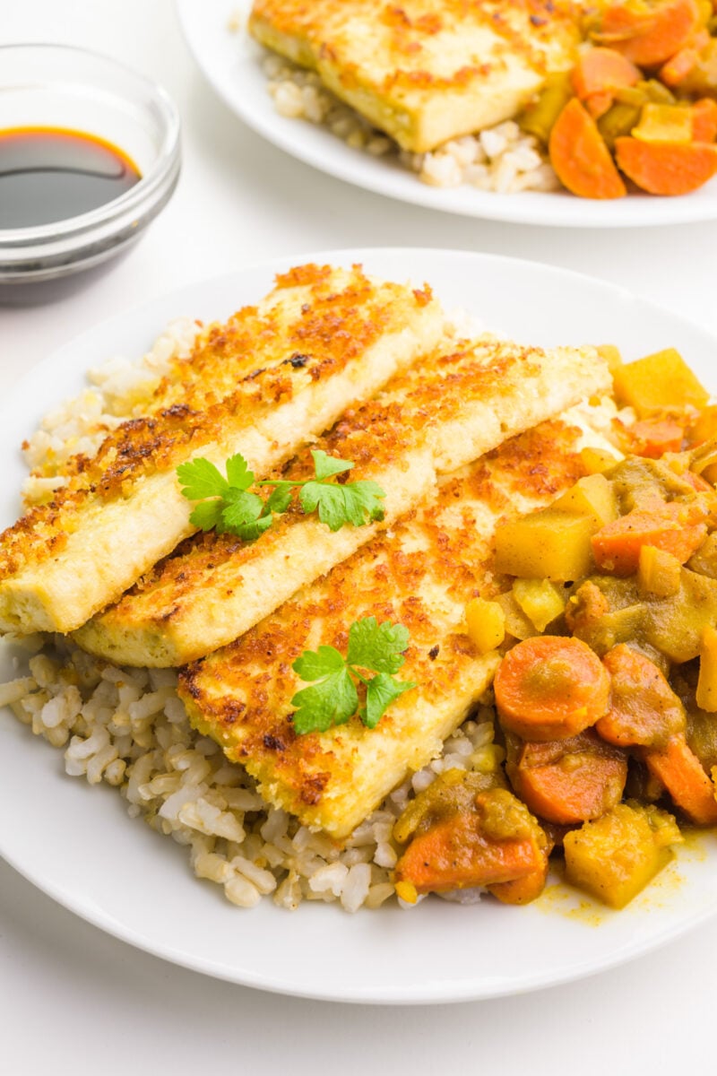 A plate has rice, fried tofu, and vegetarian katsu curry. There's a small bowl of soy sauce and another plate behind it.
