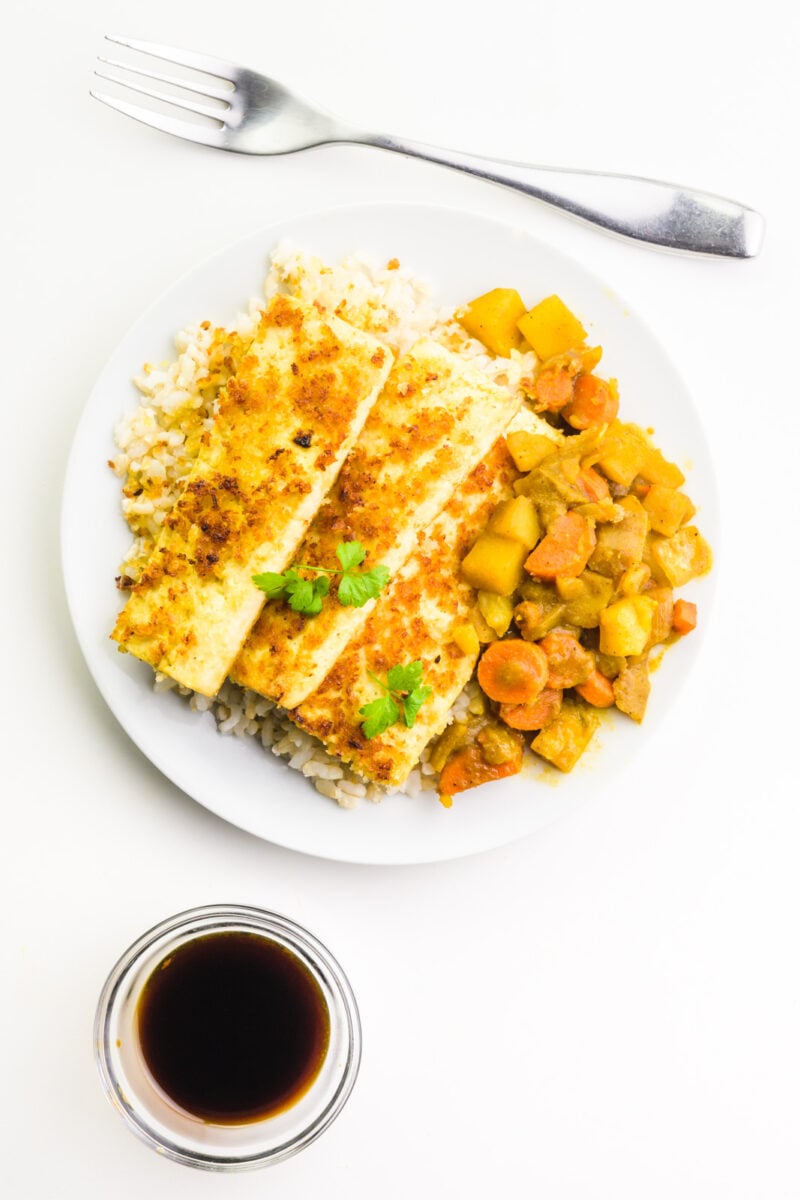 Looking down on a plate full of vegetarian katsu curry with a bowl of soy sauce on one side and a fork on the other side.