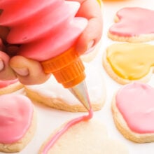 A hand holds a frosting bottle and is outlining a sugar cookie in pink frosting. there are more cookies and frosting behind it.