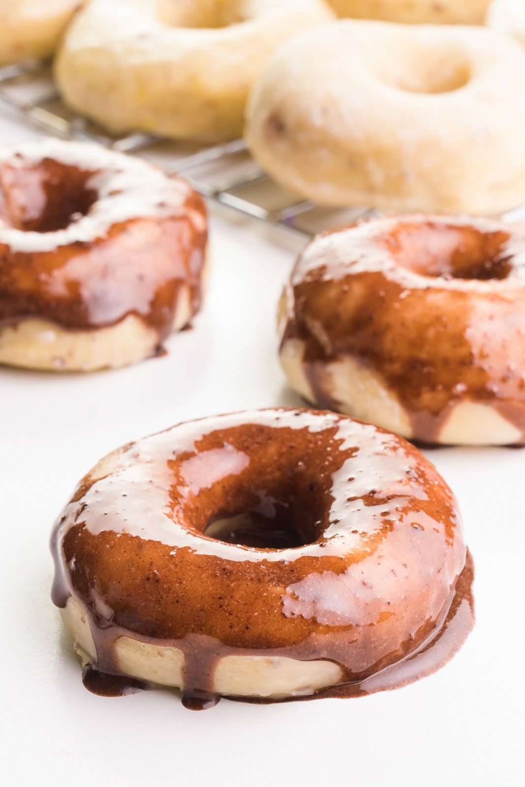 A vegan yeast donut has chocolate glaze on top and is sitting in front of two more donuts with chocolate glaze. There's a wire rack with more donuts in the background.