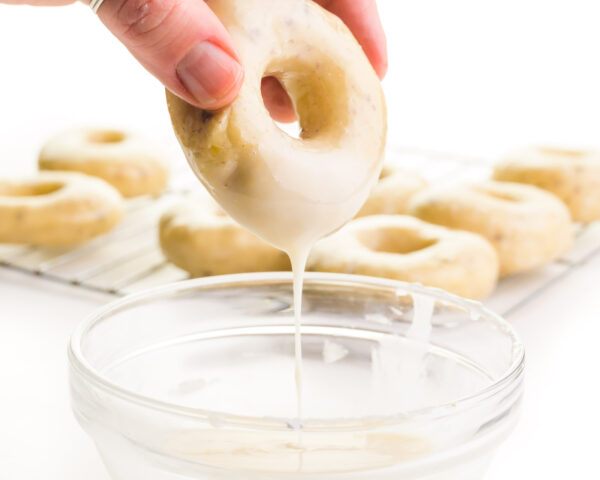 A hand holds a donut, dipping it in vanilla glaze. There are donuts on a wire rack in the background.