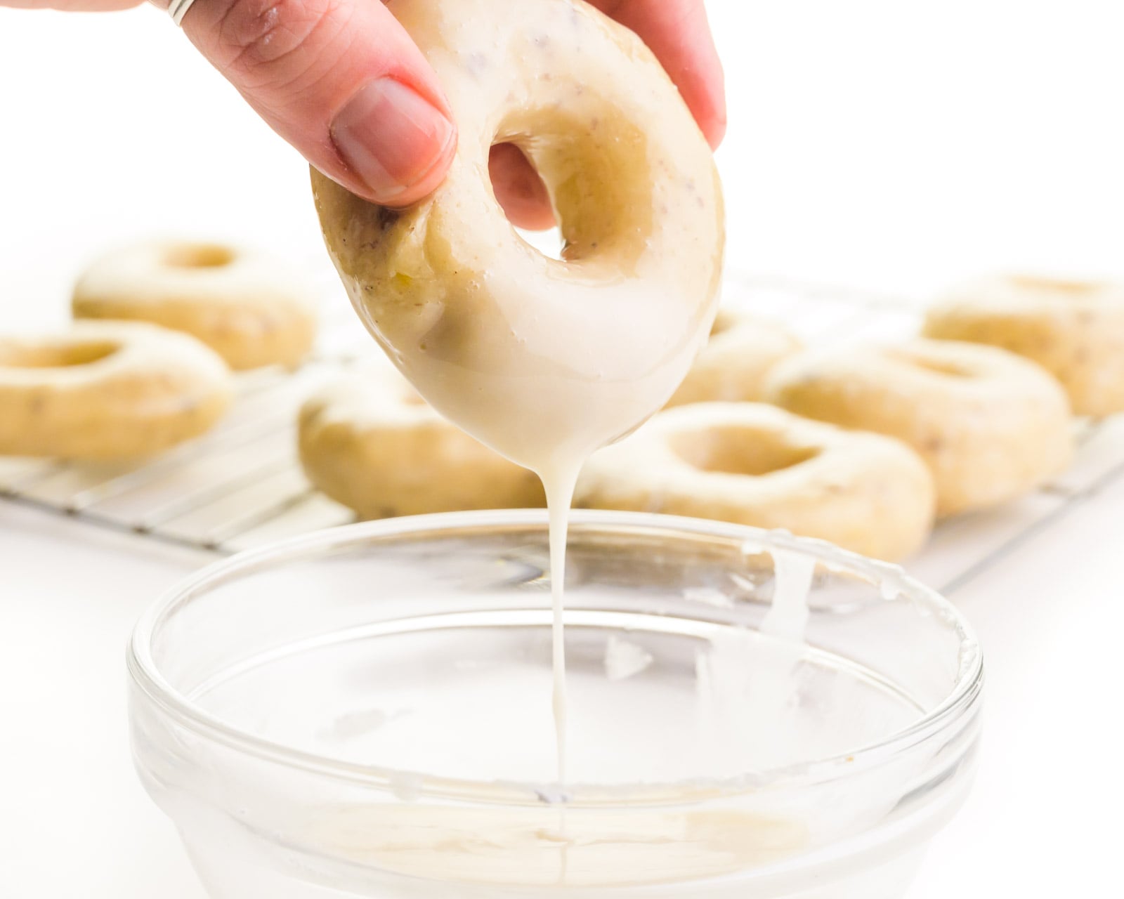 A hand holds a donut, dipping it in vanilla glaze. There are donuts on a wire rack in the background.