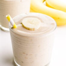 A glass holds some healthy banana shake with a yellow straw on the side. There's another glass in the background and two bananas.