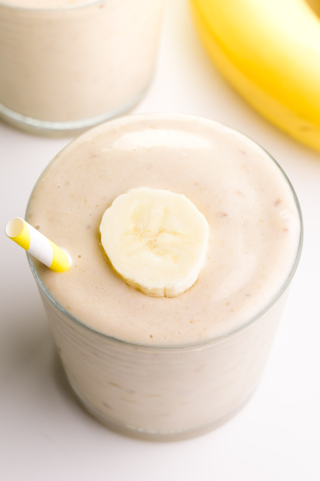 Looking down on a glass with banana smoothy. There's a banana slice on top and a yellow striped straw. There's another glass and a banana in the background.