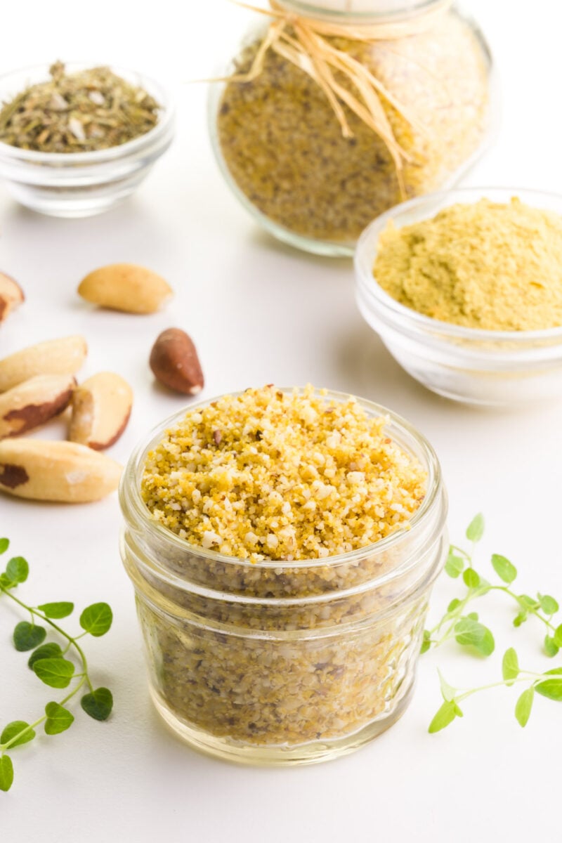 A small glass container of Brazil nut Parmesan has herbs around it. There are Brazil nuts and bowls of ingredients in the background.