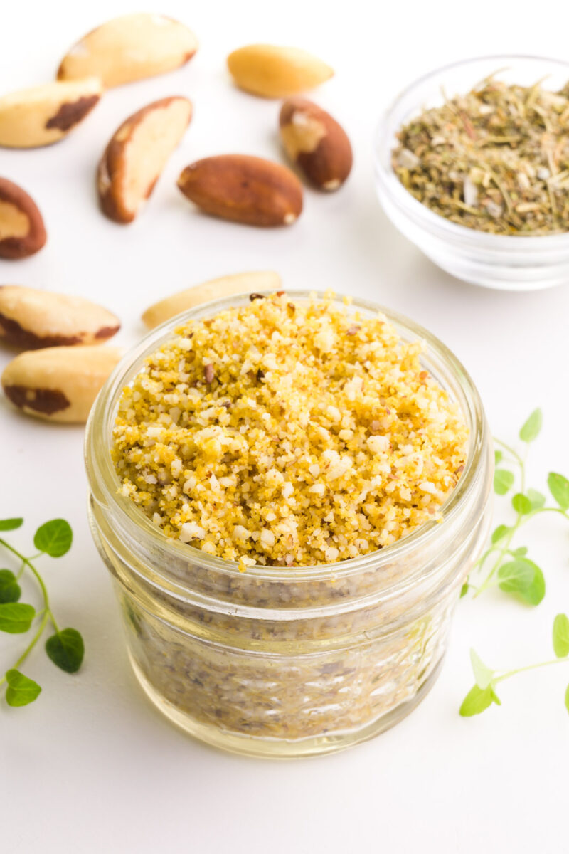 Looking down on a glass container with Brazil nut parmesan in it. There are herbs and Brazil nuts around it.