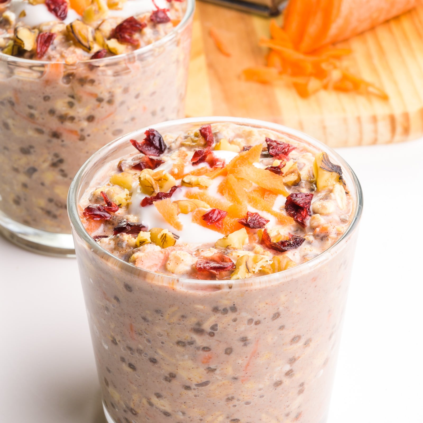 A glass holds carrot cake overnight oats with another glass behind it. There are carrots in the background.