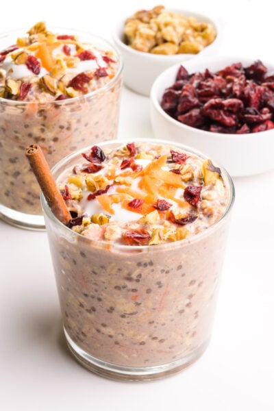 A small glass is full of carrot cake overnight oats and a cinnamon stick. There's another glass of it in the background, and a bowl of dried cranberries and walnuts.