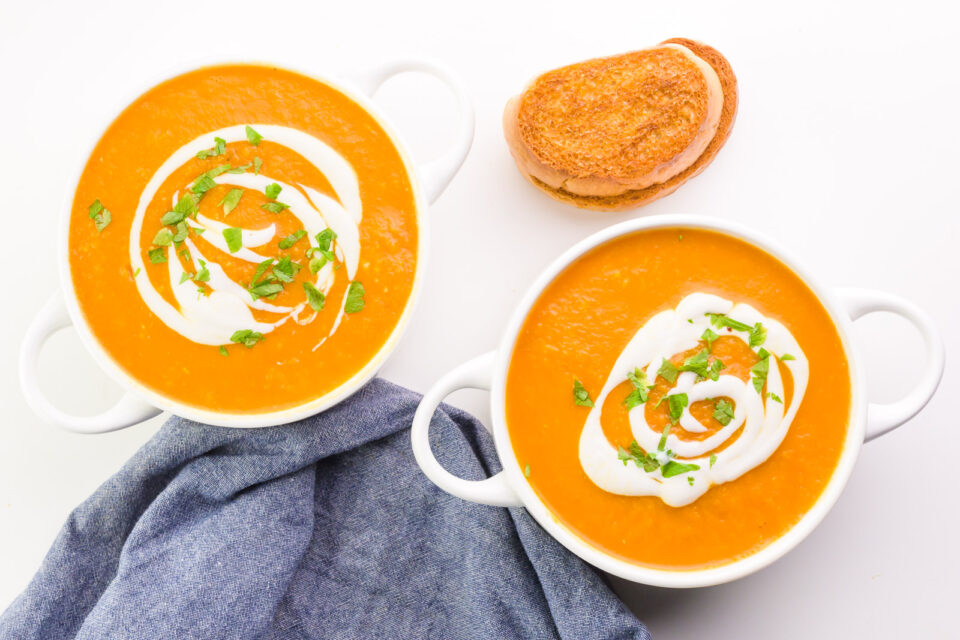 Looking down on two bowls of carrot soup sitting next to toast. There's a blue kitchen towel between the two bowls.