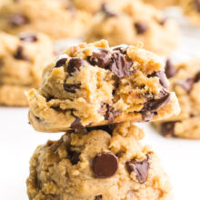 Two chunky monkey cookies are stacked with a bite taken out of the top cookie. There are more cookies in the background.