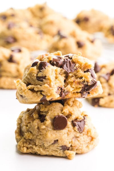 Two chunky monkey cookies are stacked with a bite taken out of the top cookie. There are more cookies in the background.