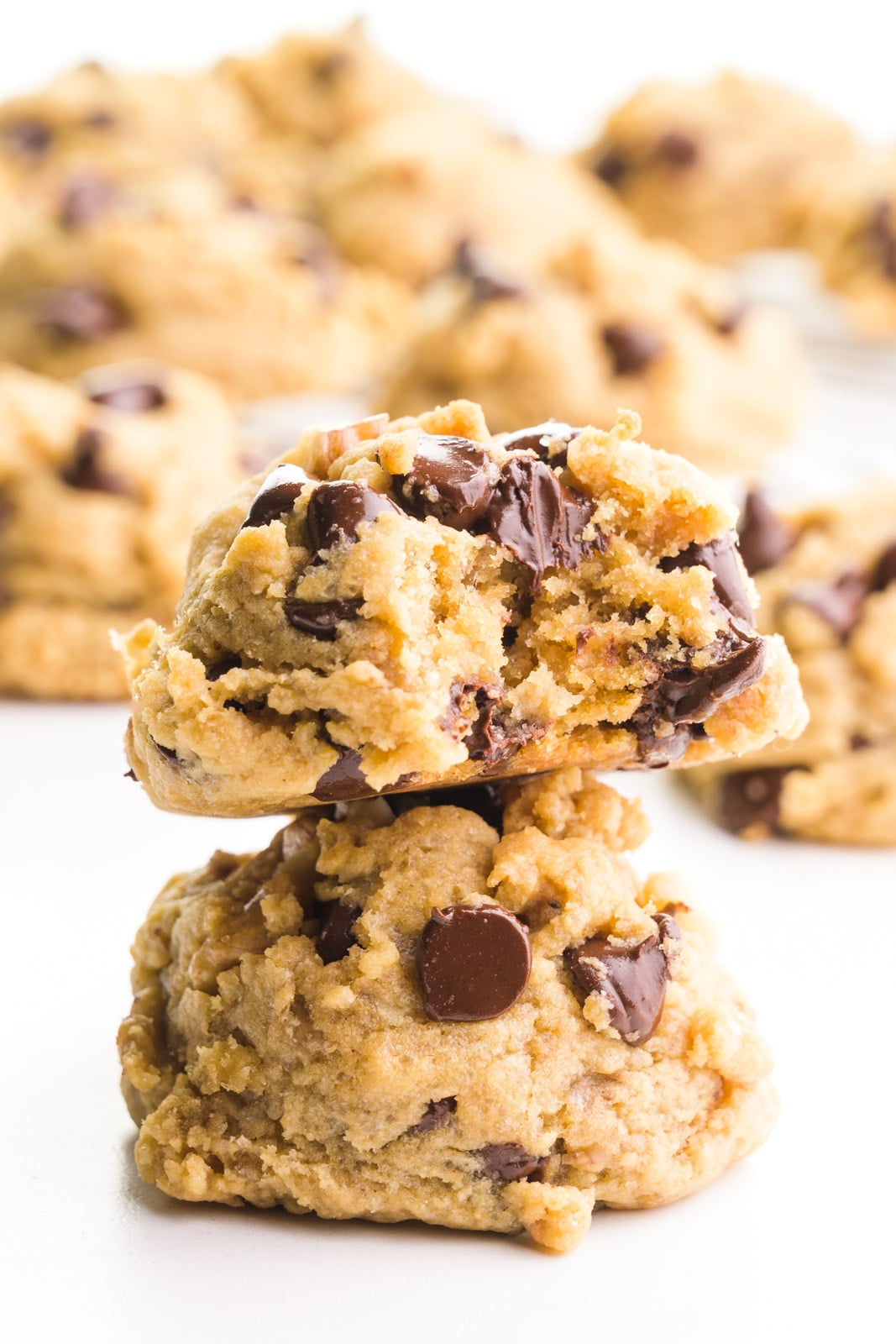 Two chunky monkey cookies are stacked with a bite taken out of the top cookie. There are more cookies in the background.