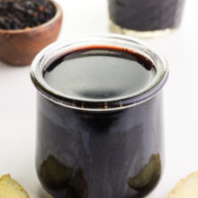 A small glass jar holds elderberry syrup. There are slices of fresh ginger in front of it. Behind it is a bowl of dried elderberries and more syrup.