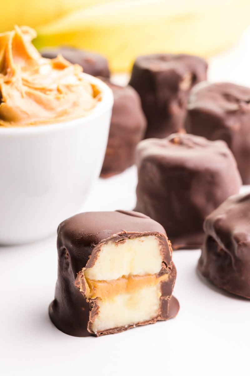 A frozen banana bite is cut in half, showing layers of banana slices and peanut butter. There are more of the treats in the background, along with a banana and a bowl of peanut butter.