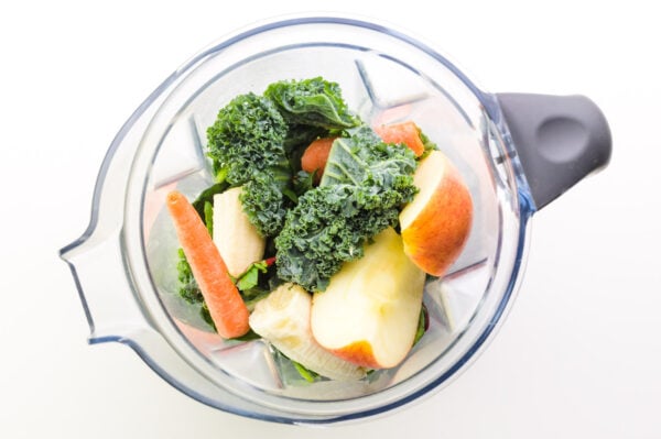 Looking down on a blender full of greens, chopped apples, bananas and carrots.