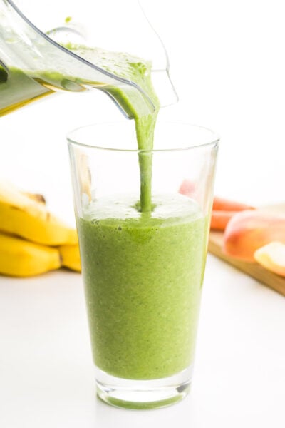 A blender is pouring a smoothie mixture into a tall glass. There are bananas in the background and a cutting board with apples on it.