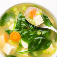 Looking down on a bowl of soup with a spoon in it. There are ingredients like greens, carrots, tofu, and green onions on top.