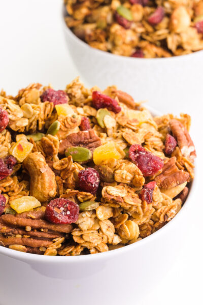 A bowl of peanut butter granola features dried cranberries, nuts, and more. There's another bowl with granola behind it.