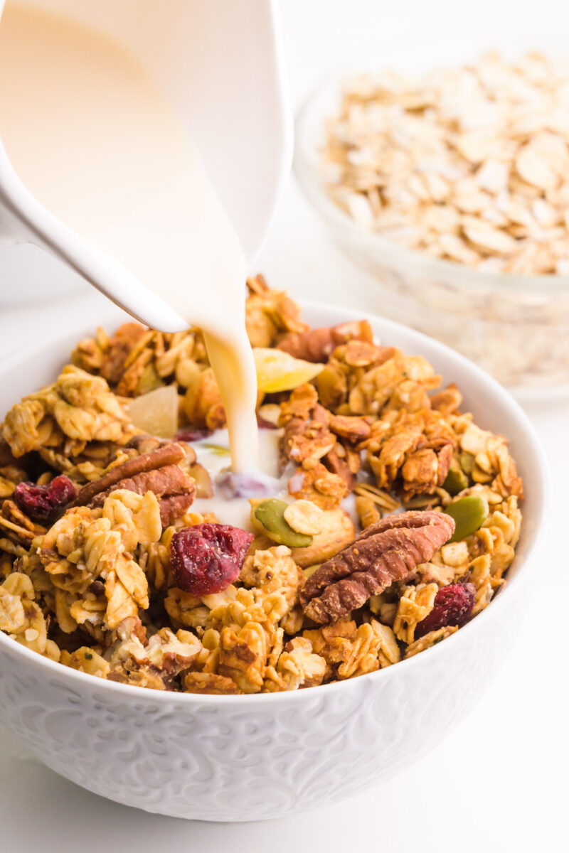 Milk is being poured into a bowl of healthy granola. There's another bowl with oats in the background.