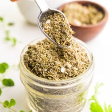 A spoon holds seasoning over a bowl of more seasoning. There are fresh herbs and bowls with more seasoning behind it.