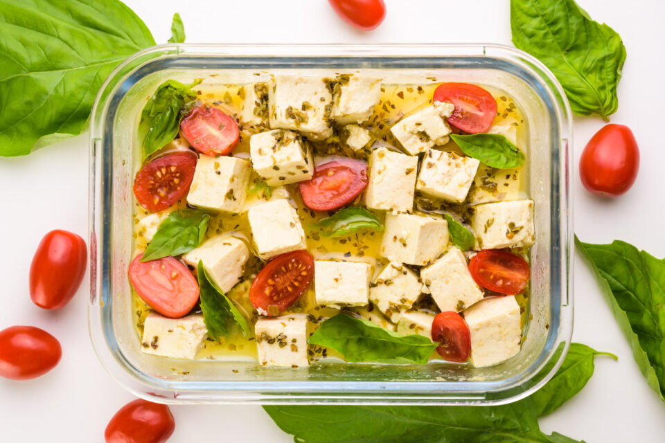 Looking down on a glass container full of tofu, cherry tomatoes, and basil leaves. There are more basil and tomatoes around the container.