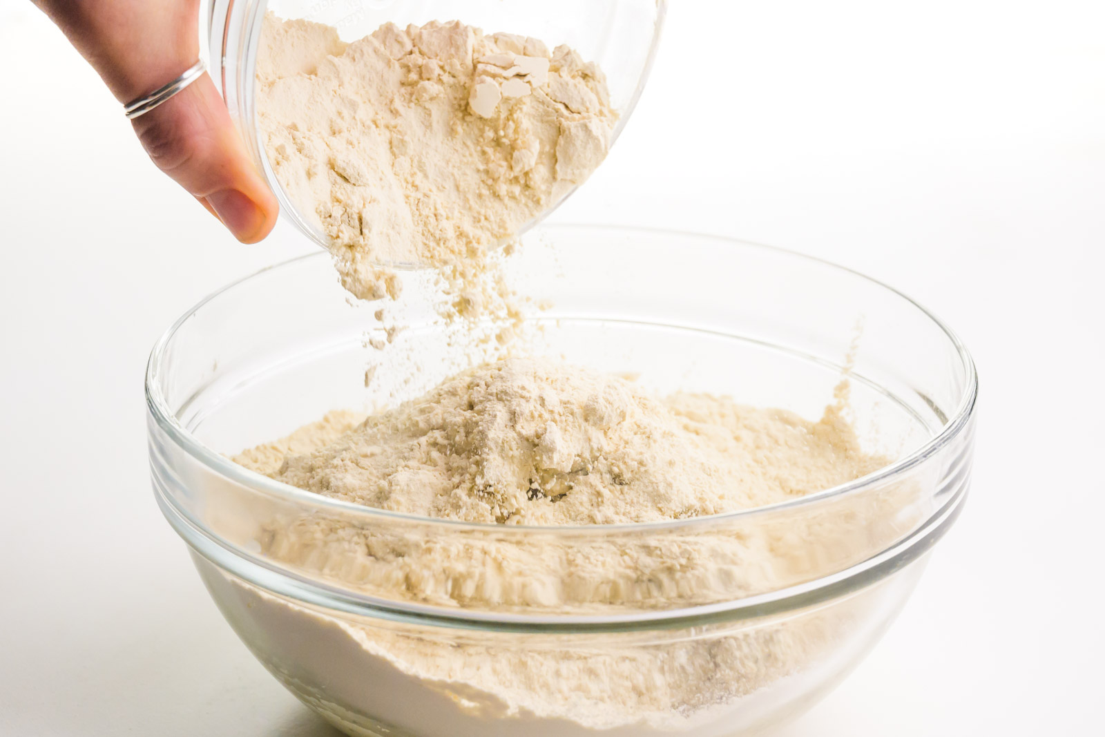 A hand holds a bowl, pouring a flour mixture over a wet mixture.