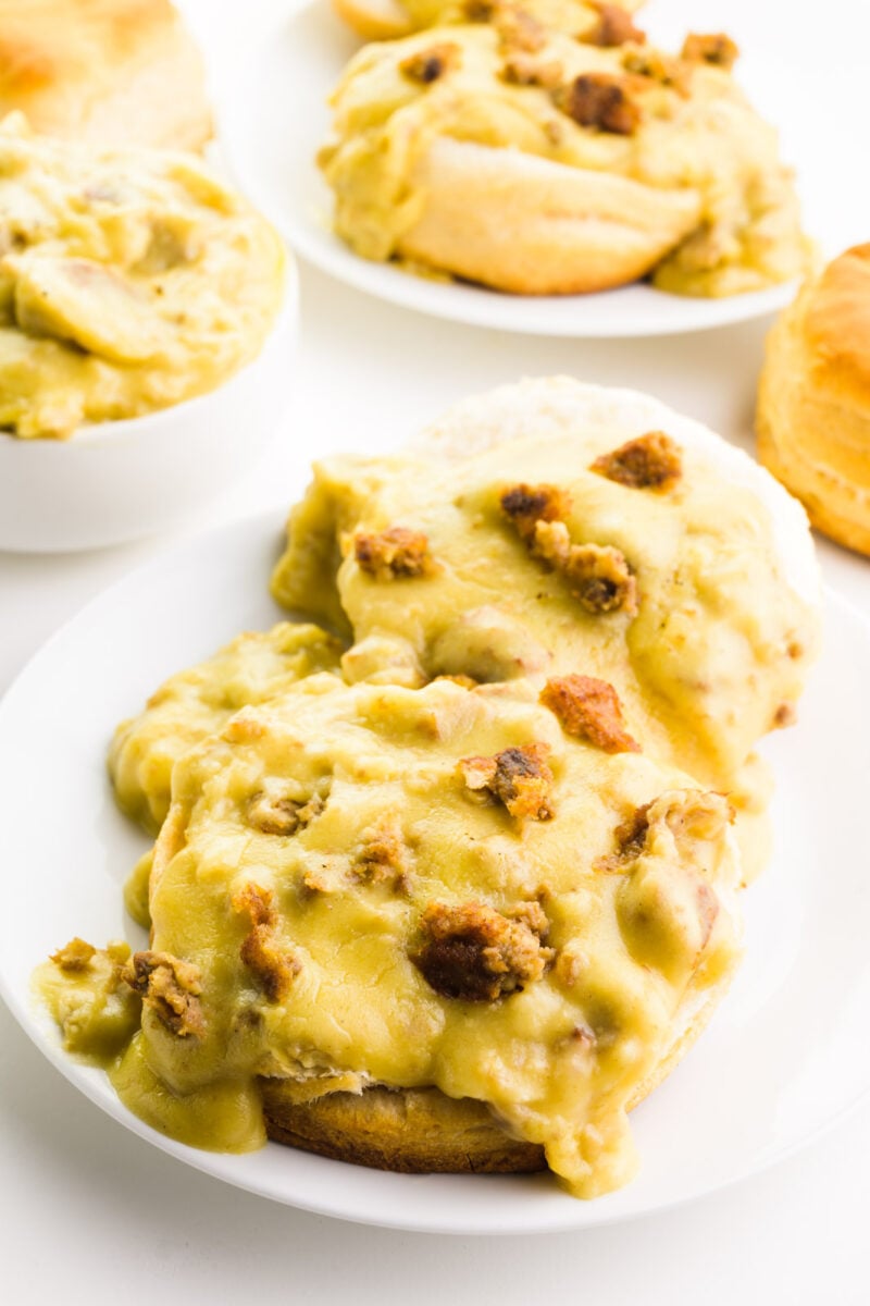 A plates holds vegan biscuits and gravy. There's a bowl with more gravy behind it along with biscuits and another plate of the dish.