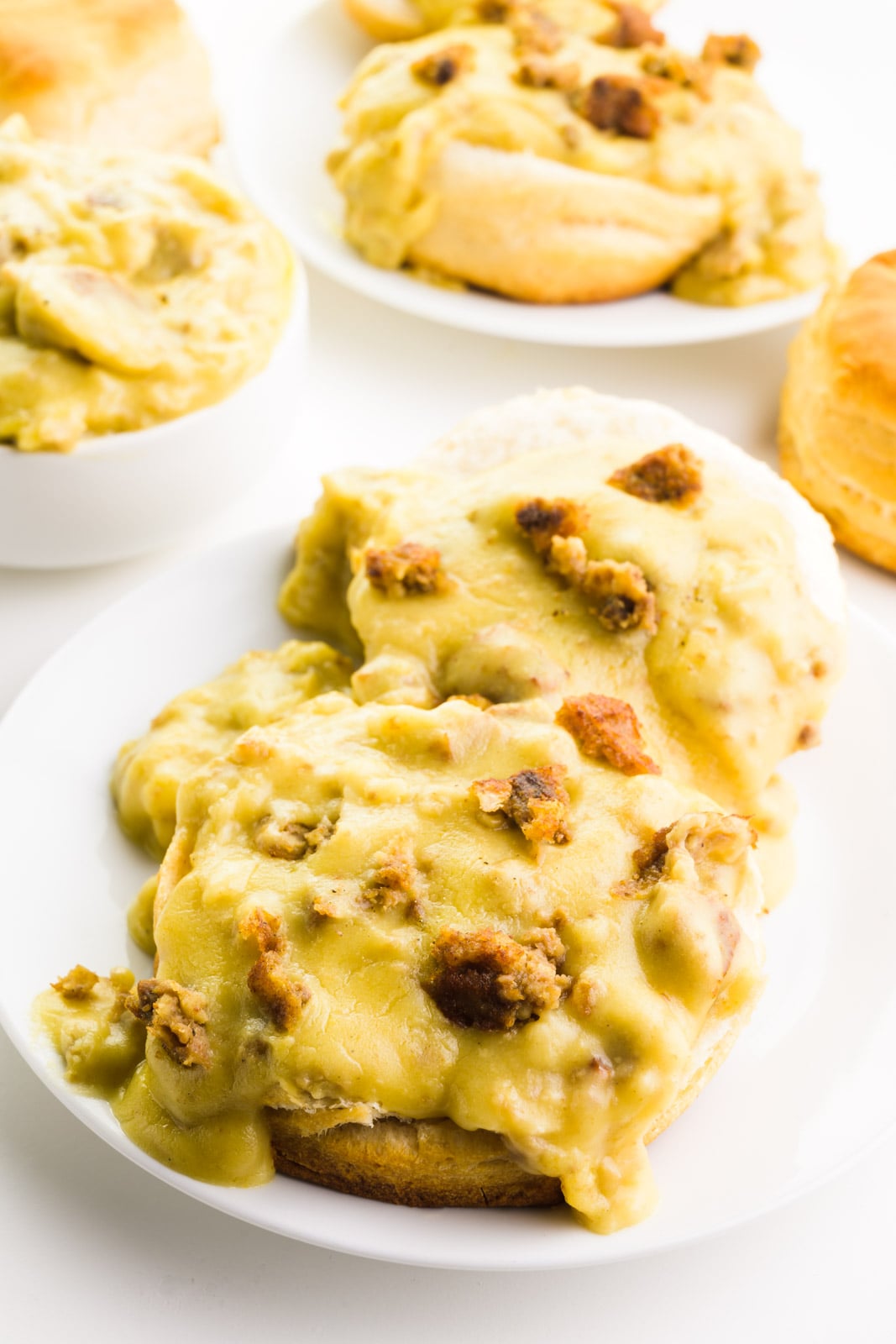 A plates holds vegan biscuits and gravy. There's a bowl with more gravy behind it along with biscuits and another plate of the dish.
