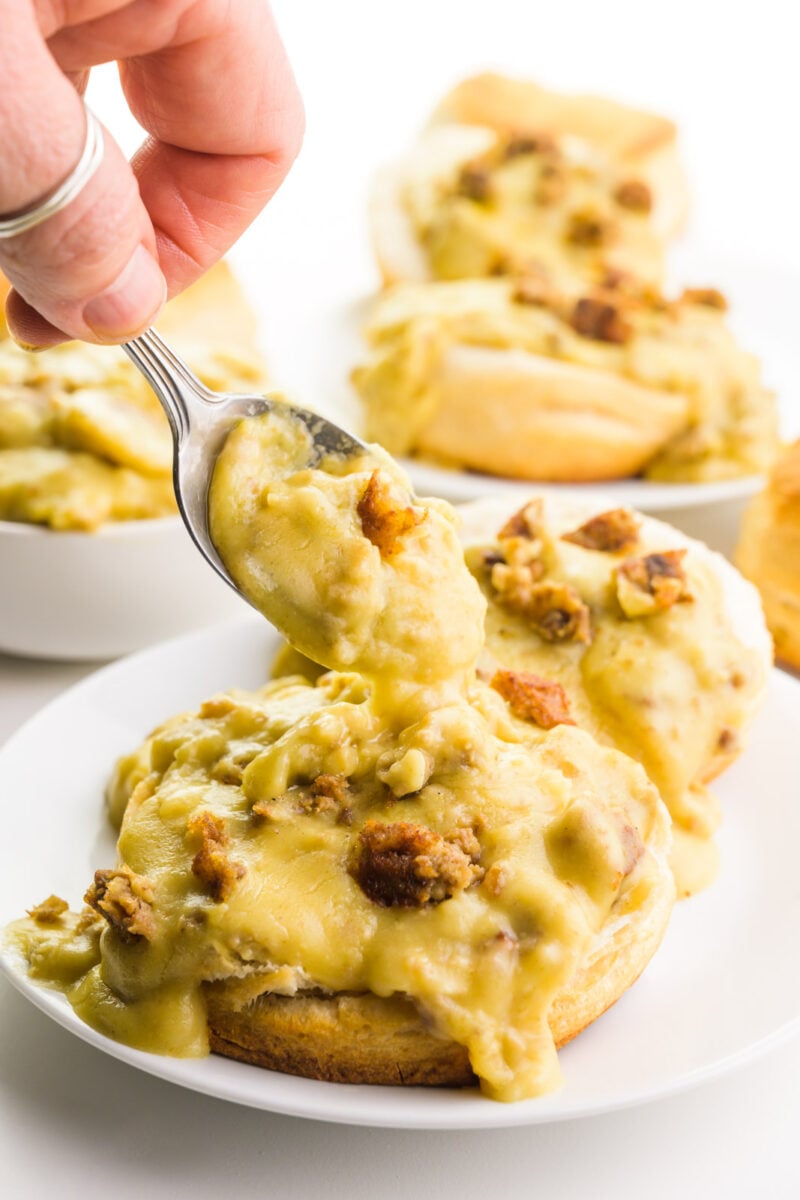 A spoon is dishing gravy onto a plate with biscuits and gravy. There are more ingredients behind the plate.