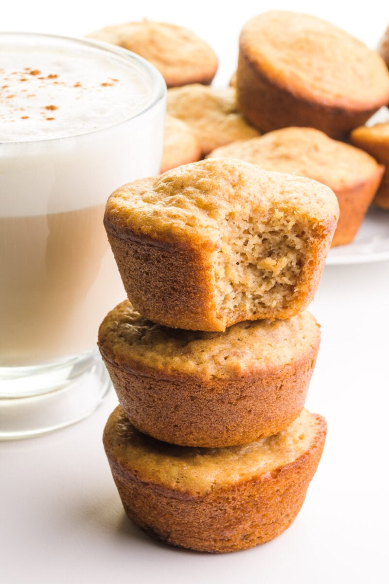 A stack of vegan bran muffins shows a bite taken out of the to one. There's a mug of tea with frothy milk on top and more muffins in the background.