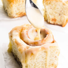 Three vegan cinnamon rolls are on a counter. A hand holds a spoon drizzling frosting over the top of one of them.