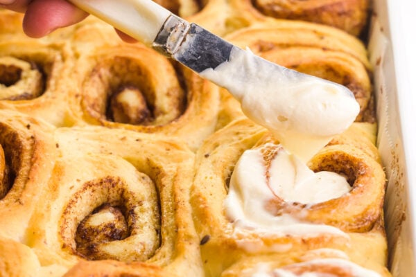 A butter knife is used to drizzle frosting over freshly baked cinnamon rolls.