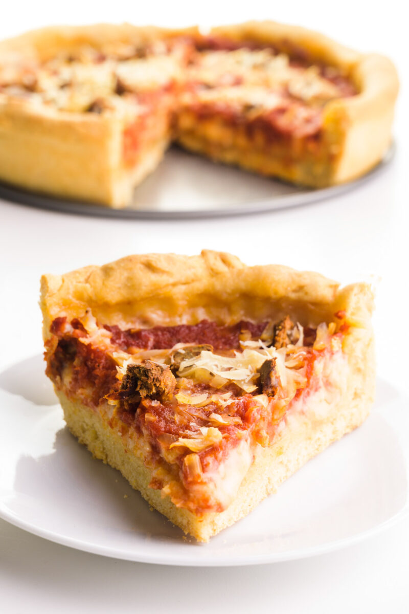 A slice of vegan pizza sits on a plate with the rest of the pizza on a tray in the background.