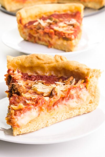 A slice of vegan deep-dish pizza sit on a plate. Another slice is on a plate behind it with the rest of the pizza.