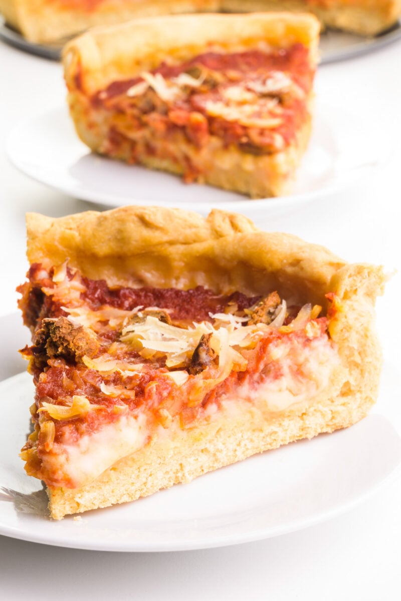 A slice of vegan deep-dish pizza sit on a plate. Another slice is on a plate behind it with the rest of the pizza.