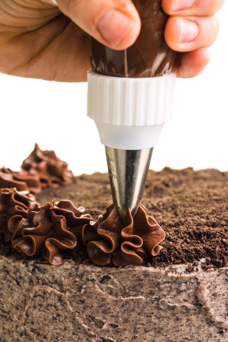 A hand holds a piping bag adding dollops of vegan ganache to a chocolate cake.