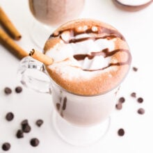 Looking down on a mug full of hot cocoa with whipped cream and chocolate syrup on top. There's a cinnamon stick in the mug and more beside. There are chocolate chips around the mug.