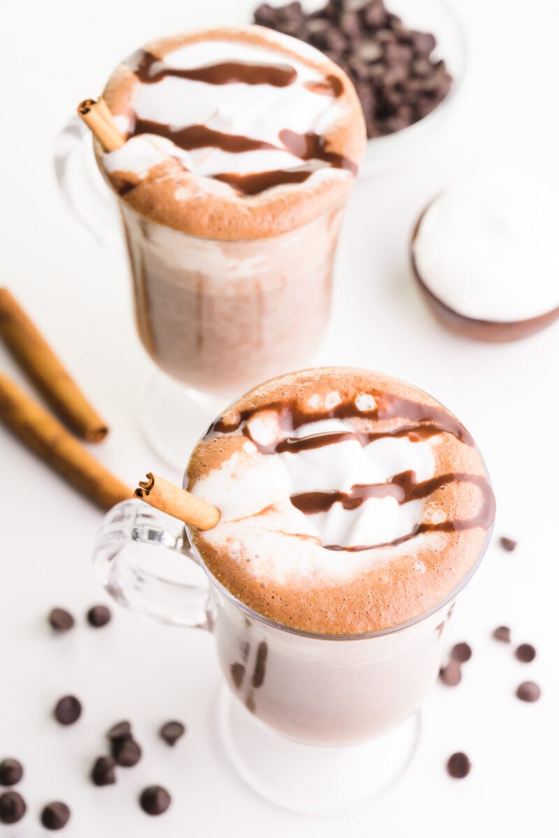 Lookin down on two glass mugs with vegan hot cocoa. They have cream and chocolate syrup on top. There are cinnamon sticks in both mugs. There are chocolate chips and cinnamon sticks around the mugs and a bowl of whipped cream and chocolate chips.