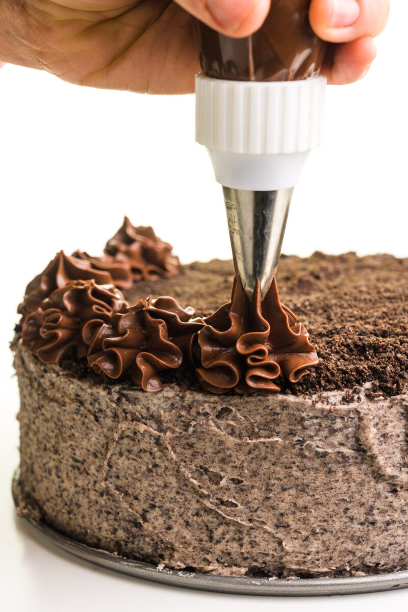A hand holds a piping bag, adding chocolate ganache stars on top.